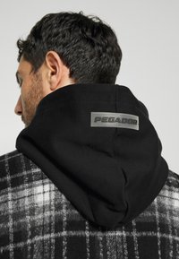Homme aux cheveux courts et foncés portant un vêtement à capuche noir étiqueté "PEGADOR" par-dessus une veste à carreaux noir et blanc, vu de dos.