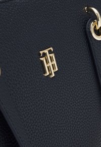 Marinblå strukturerad läderhandväska med en guldtonad logotypdetalj och metallhardware. Närbild visar materialet och designens detaljer.