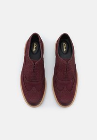 Une paire de chaussures brogues en suede bordeaux avec des semelles brunes et des semelles intérieures noires, photographiées de dessus sur un fond blanc.