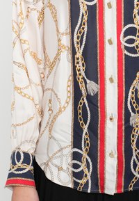 Bijela bluza s uzorkom zlatnih lanaca i uže, mornarsko plave i crvene okomite pruge, dugi rukavi, šiljati ovratnik i ukrasni gumbi.
