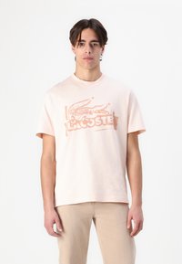 Helles rosa T-Shirt mit Rundhalsausschnitt, das ein strukturiertes Krokodil-Logo und den "Lacoste"-Schriftzug in einer kontrastierenden Farbe zeigt. Weiches Baumwollmaterial, entspannter Schnitt.