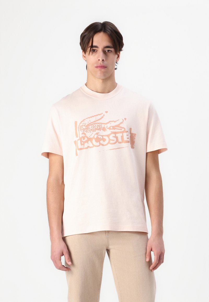 Helles rosa T-Shirt mit Rundhalsausschnitt, das ein strukturiertes Krokodil-Logo und den "Lacoste"-Schriftzug in einer kontrastierenden Farbe zeigt. Weiches Baumwollmaterial, entspannter Schnitt.