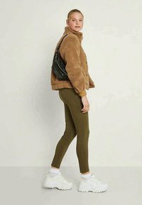 Jovem mulher vestindo uma jaqueta de teddy castanha, leggings oliva, ténis brancos e uma mala de ombro preta, de pé de lado contra um fundo simples.