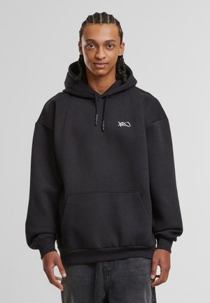 Schwarzer Kapuzenpullover mit einer vorderen Kängurutasche, gerippten Bündchen und einem kleinen weißen Logo auf der Brust. Hergestellt aus weichem, dickem Stoff.