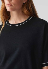 Schwarzer Strickpullover mit Rundhalsausschnitt, der einen kontrastierenden weißen Saum entlang des Halsausschnitts und der Ärmelenden aufweist. Weiche Textur.