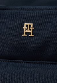 Φόντο από υφασμάτινη ύφανση ναυτικού μπλε χρώματος με χρυσαφένιο λογότυπο που απεικονίζει διαπλεγμένα γράμματα "H" και "T", παρουσιάζοντας κομψό σχεδιασμό και υφή επιφάνειας.