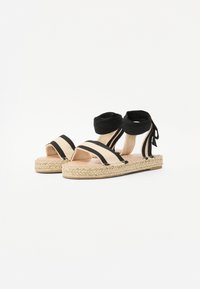 Sandale espadrile negre și bej cu vârf deschis și legături la gleznă, având o talpă din iută țesută și un design din țesătură cu dungi contrastante.