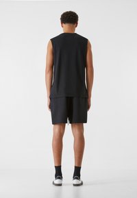 Schwarzes ärmelloses Oberteil mit lockerer Passform, kombiniert mit schwarzen Shorts. Model trägt weiße Schuhe und schwarze Socken. Einfaches, minimalistisches Design.
