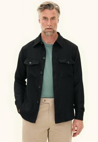 Black button-up shirt jacket with two chest pockets and a textured finish, worn over a light green crew neck sweater, paired with beige pants.