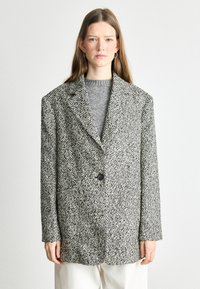 Textureeritud must-valge tweed bleiser ühe nupu kinnitusega, laiade krae ja rõhutatult l�õdvema lõikega, mille alla on sobitatud hall sviiter.