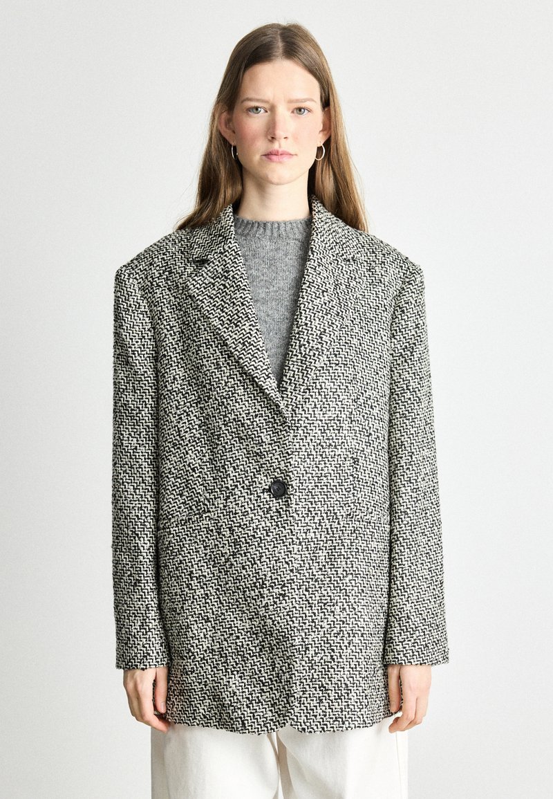 Textureeritud must-valge tweed bleiser ühe nupu kinnitusega, laiade krae ja rõhutatult lõdvema lõikega, mille alla on sobitatud hall sviiter.