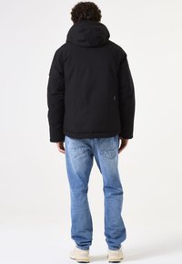 Chaqueta acolchada negra con capucha, con una textura suave y un branding mínimo. Combinada con unos vaqueros de mezclilla azul claro y zapatillas beige.