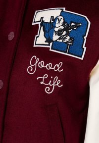 Burgundowa kurtka varsity z mieszanki wełny, z niebiesko-białym haftowanym logo i napisem "Good Life" w białym kursywie z przodu.