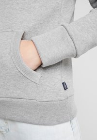 Sweat-shirt gris avec poche kangourou, poignets et ourlet côtelés. Le tissu présente une légère texture ; petite étiquette logo visible sur le côté.