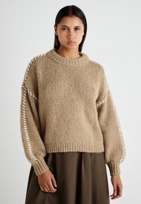 Beige strikket genser med rund hals, strukturert stoff og åpne sømmer langs ermene. Kombinert med en mørk skjørt.
