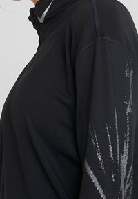 Czarna sportowa bluzka z długim rękawem i zamkiem na ćwierć, z logo Nike na kołnierzu oraz odblaskowym punktowym wzorem na rękawie.