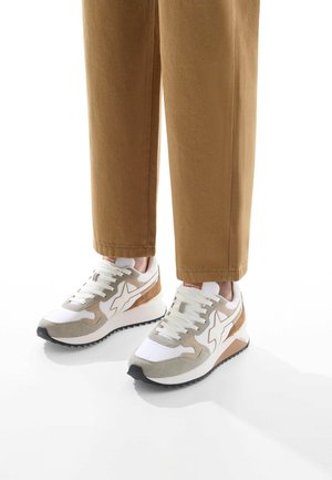 Zapatillas con un upper de textil blanco y beige, que presentan un logo prominente, detalles en ante y una suela de goma negra.