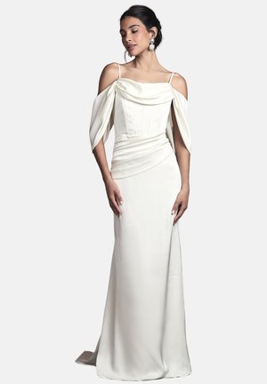 Donna che indossa un elegante abito bianco con spalle scoperte, maniche drappeggiate e spalline sottili, in piedi con le mani rilassate lungo i fianchi.