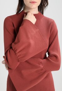 Rippengestricktes Langarmoberteil in Ziegelrot mit einem hohen Halsausschnitt und ausgestellten Ärmeln mit elastischen Bündchen. Weicher, strukturierter Stoff.