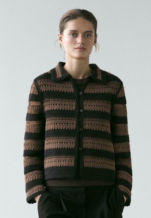 Donna che indossa un cardigan all'uncinetto a righe nere e marroni con bottoni e colletto, in piedi con le mani nelle tasche contro uno sfondo semplice.