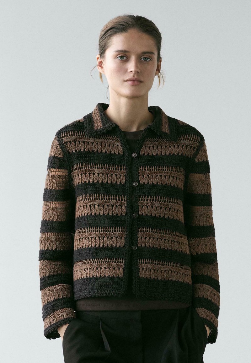 Frau trägt eine schwarz-braun gestreifte, gehäkelte Strickjacke mit Knöpfen und Kragen, steht mit den Händen in den Taschen vor einem schlichten Hintergrund.