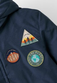 Jūras spēku jaka ar trim auduma uzlīmes: trīsstūru zaļā un oranžā, apaļā oranžā ar vārdu "DROŠS," un aplīša zilā ar vārdu "MĪLI PLANETU."