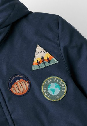 Námornícky kabát s tromi textilnými nášivkami: trojuholníková zelená a oranžová, okrúhla oranžová s nápisom "BRAVE" a kruhová modrá s nápisom "LOVE THE PLANET."