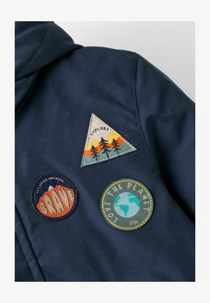 Tumesinine jakk kolme riidepatšiga: kolmnurkne roheline ja oranž, ümmargune oranž, millele on kirjutatud "BRAVE", ja ümmargune sinine, millele on kirjutatud "LOVE THE PLANET."