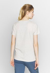Vrouw met een blonde paardenstaart, die zijwaarts kijkt, gekleed in een lichtbeige T-shirt met korte mouwen en blauwe broek met chevronpatronen.