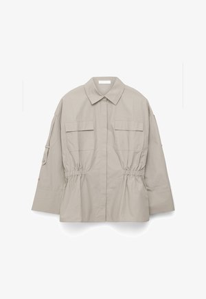 Beige lange mouw jas met puntkraag, twee voorzakken met klep, een getailleerde taille en knoopdetail aan de mouwen, plat liggend op een witte achtergrond.