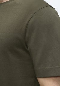 Gros plan d'une personne portant un t-shirt à manches courtes vert olive montrant la texture du tissu et le haut du bras.