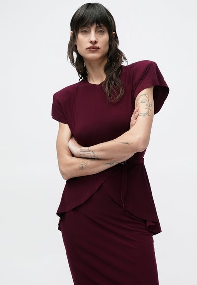 Osoba o ciemnych włosach sięgających ramion i tatuażach na ramionach, ubrana w dopasowaną, bordową sukienkę z krótkim rękawem, stojąca ze skrzyżowanymi ramionami.