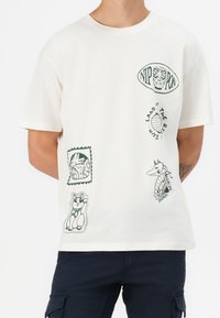 Bílé tričko s zelenými japonským tématickými potisky, včetně panenky Daruma, lucerny s nápisem "Slunce vychází", lišky s nápisem "Vyrobeno v Japonsku" a mávající kočky.