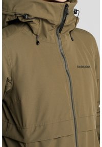 Veste verte olive, résistante à l'eau, avec un col haut, un zip à l'avant, une capuche ajustable et un détail logo noir. Présente une texture lisse.