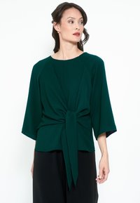Donna che indossa una blusa verde scuro con nodo frontale e maniche ampie, che guarda verso sinistra su uno sfondo bianco.