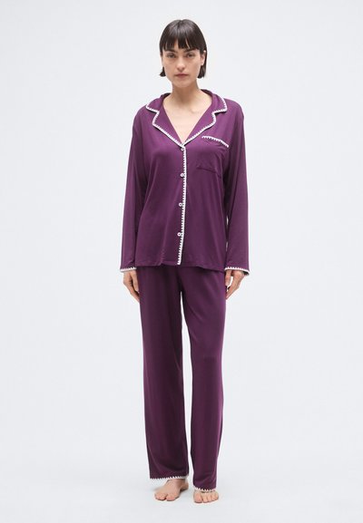 Ensemble de pyjama violet comprenant un haut à boutons avec une poche poitrine et un bord blanc, associé à un pantalon long, tous deux fabriqués dans un tissu doux et texturé.