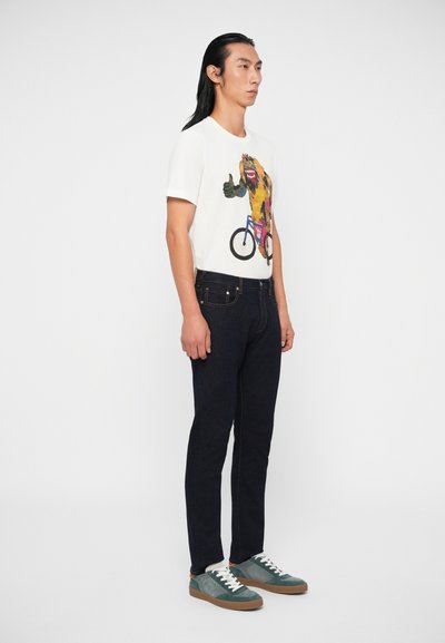 T-shirt gráfica branca com um design colorido de animais, combinada com jeans escuros e ténis verdes com atacadores brancos e solas castanhas.