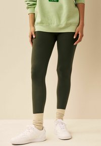 Olivenfarvede leggings med en glat tekstur, tætsiddende design og ankelmanchetter. Kombineret med hvide sneakers og en lysegrøn sweatshirt.