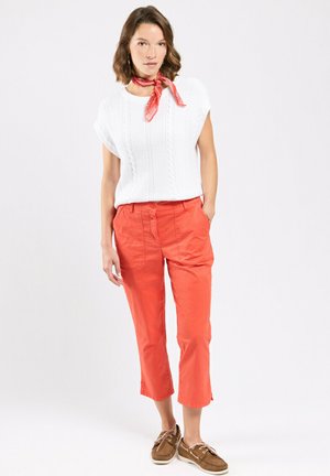 Femme portant un haut sans manches en tricot blanc, un pantalon rouge raccourci, un foulard rouge autour du cou et des chaussures bateau marron, debout devant un fond blanc.