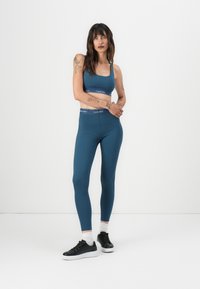 Tirkizni bezšavne sportske grudnjake i tajice s Calvin Klein logotipom na struku. Model nosi crne tenisice s bijelim potplatima i čarape do gležnja.