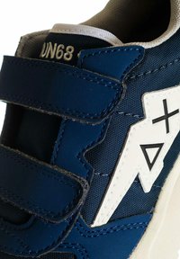 Sneaker blu navy con strap in Velcro, tomaia in tessuto testurizzato, accenti geometrici bianchi e colletto imbottito per il comfort. Branding distintivo visibile.