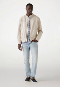 Beige jas met ritssluiting aan de voorkant, gestreept T-shirt, lichtblauwe spijkerbroek en donkere schoenen. Eenvoudig ontwerp met een casual pasvorm en zonder zichtbare patronen of logo's.