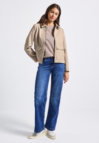 Žena stojící v béžové zipové bundě, světle hnědém tričku, modrých džínách a bílých teniskách na jednotném světlém pozadí.