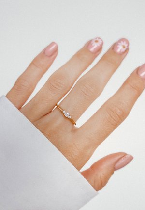 Goldring mit drei kleinen klaren Edelsteinen, auf einem schmalen Band, getragen an einer Hand mit zart rosa Nagellack und Blumenmotiven.