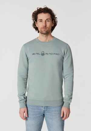 Sudadera gris claro de manga larga, con cuello redondo y un estampado gráfico "SAIL RACING" en el pecho. Tejido de textura suave. Jeans de mezclilla.