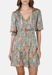 Femme portant une robe portefeuille colorée et à motifs avec des manches bouffantes et un col en V, se tenant devant un fond clair.