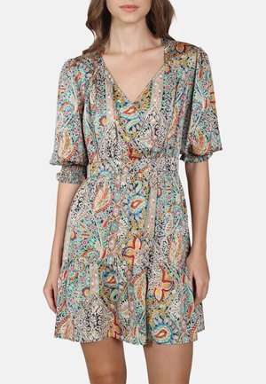 Femme portant une robe portefeuille colorée et à motifs avec des manches bouffantes et un col en V, se tenant devant un fond clair.