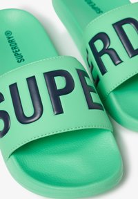 Groene schuimslippers met een gestructureerde band waarop grote marineblauwe letters het woord "SUPER" spellen. Gemaakt van synthetisch materiaal met een gladde afwerking.