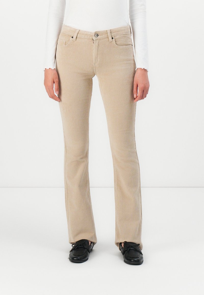 Pantalon en velours côtelé beige porté avec des chaussures noires à lacets et un haut blanc à manches longues, visible de la taille aux pieds sur fond blanc.