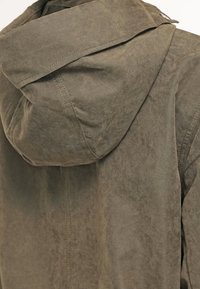 Olivově zelená bunda s kapucí vyrobená z texturované látky, viditelné švy a mírně lesklý povrch. Má uvolněný střih a skládací límec.